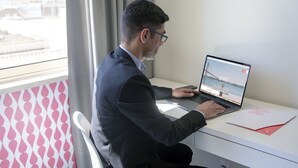 A person sitting at a desk with a laptop, a red folder, and a window with curtains. - Stay Hotel Faro Centro (Faro)