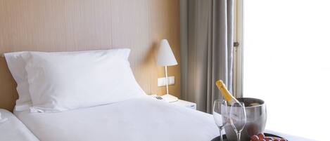 A hotel room with a neatly made bed, a bedside table with a lamp, a bucket with champagne, and a tray with grapes and glasses.