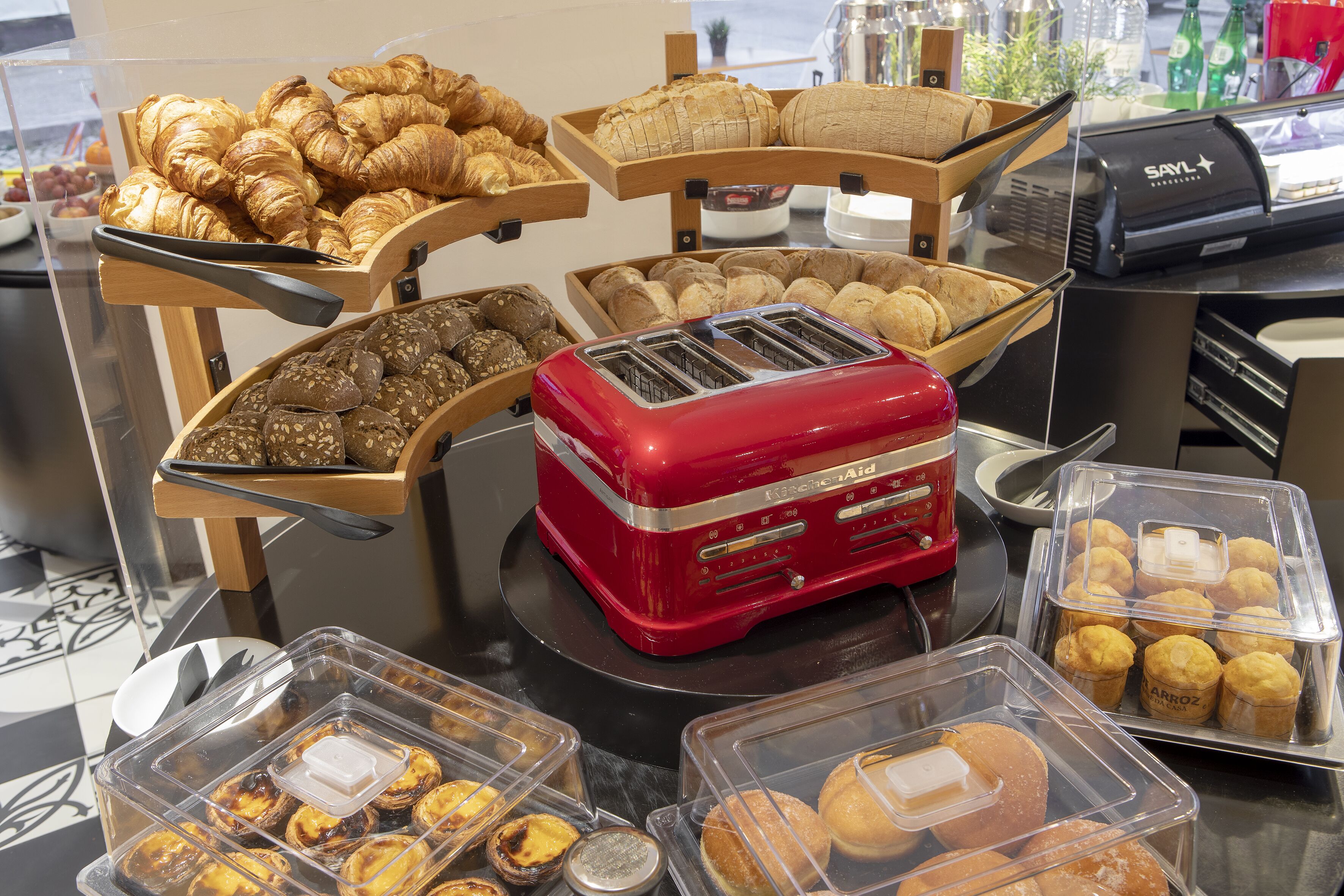 Un bufé con diversos productos horneados, como croissants, pan, muffins y pasteles, exhibidos sobre bases de madera y en recipientes transparentes.