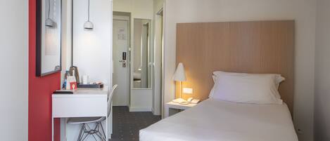 A hotel room with a bed, a desk with a chair, a lamp, and a mirror.