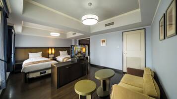 Quarto de hotel com cama, escrivaninha, duas mesas redondas, sofá e televisão.
