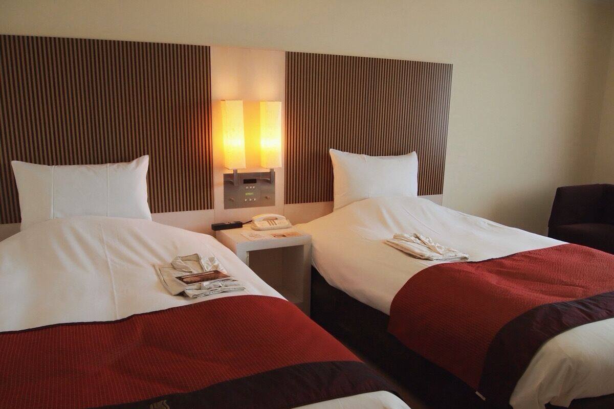 Une chambre d’hôtel avec deux lits, chacun agrémenté de draps blancs et de touches de couleur rouge. On y trouve une table de chevet avec une lampe, un téléphone et un magazine. La chambre présente un design moderne, avec un revêtement en bois sur la tête de lit.