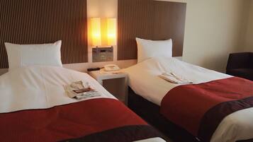 Habitación de hotel con dos camas, cada una con ropa blanca y detalles rojos, una mesita de noche con lámpara y un teléfono.