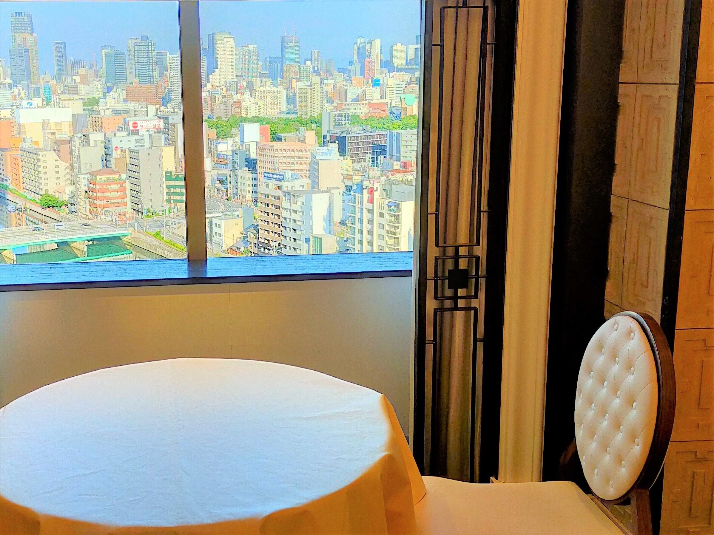 A hotel room with a large window offering a city view, a round white table, and a tufted chair.