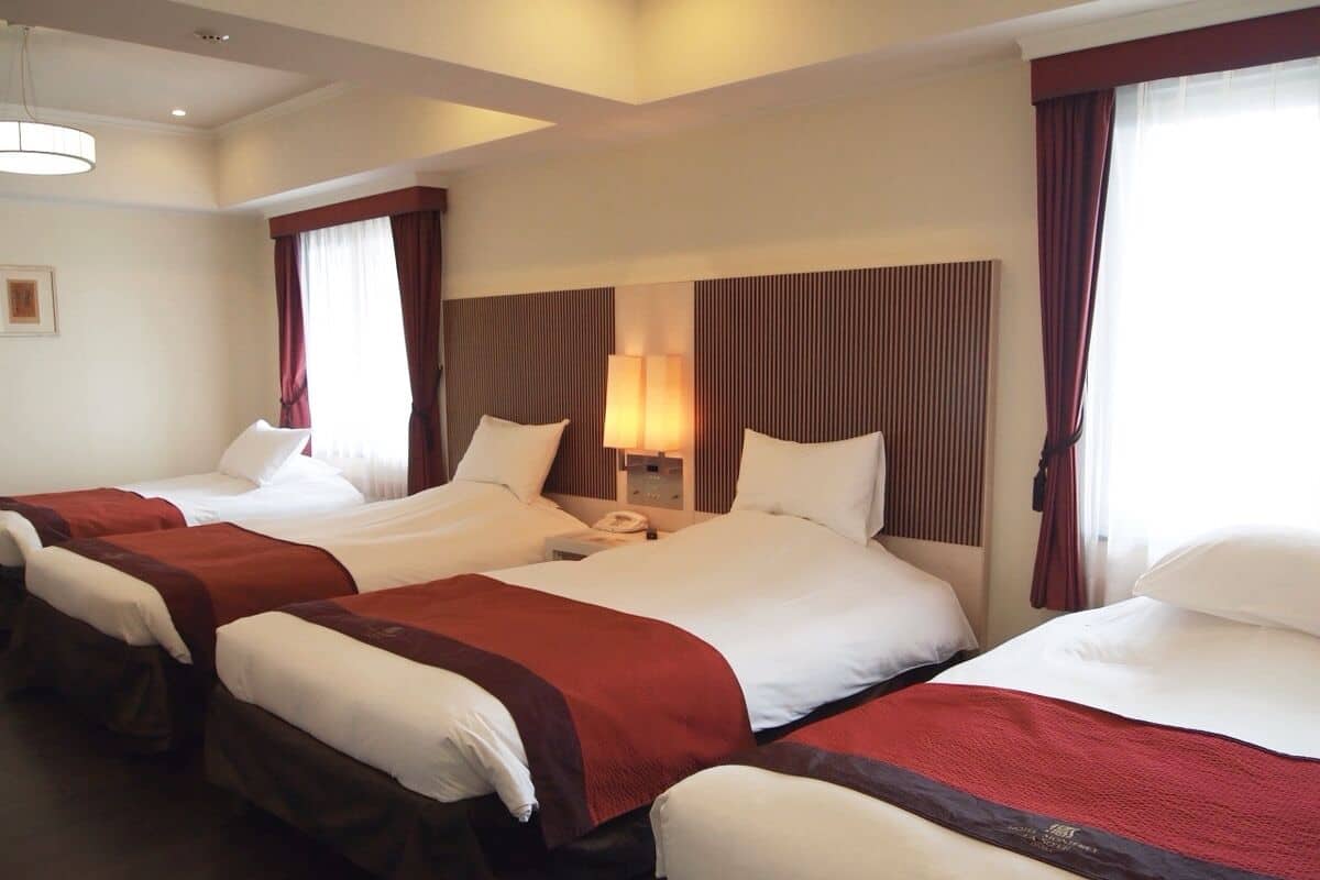 Une chambre d’hôtel avec quatre lits simples, chacun recouvert d’une couverture rouge et blanche, et une grande fenêtre avec des rideaux.