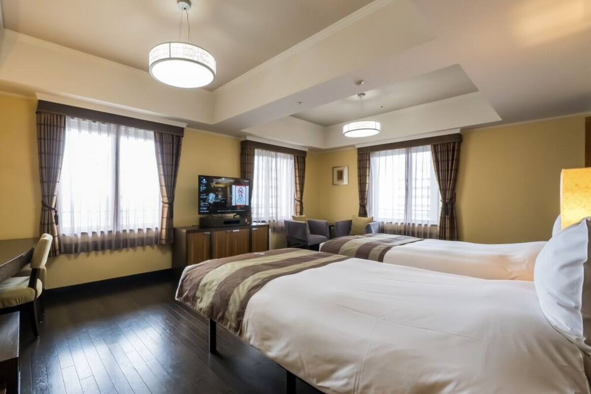 Une chambre d’hôtel avec deux lits, un bureau, une télévision et de grandes fenêtres avec des rideaux.