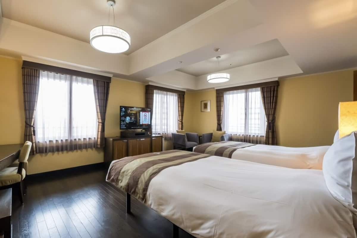 Quarto de hotel com duas camas, uma escrivaninha, uma televisão e janelas amplas com cortinas.