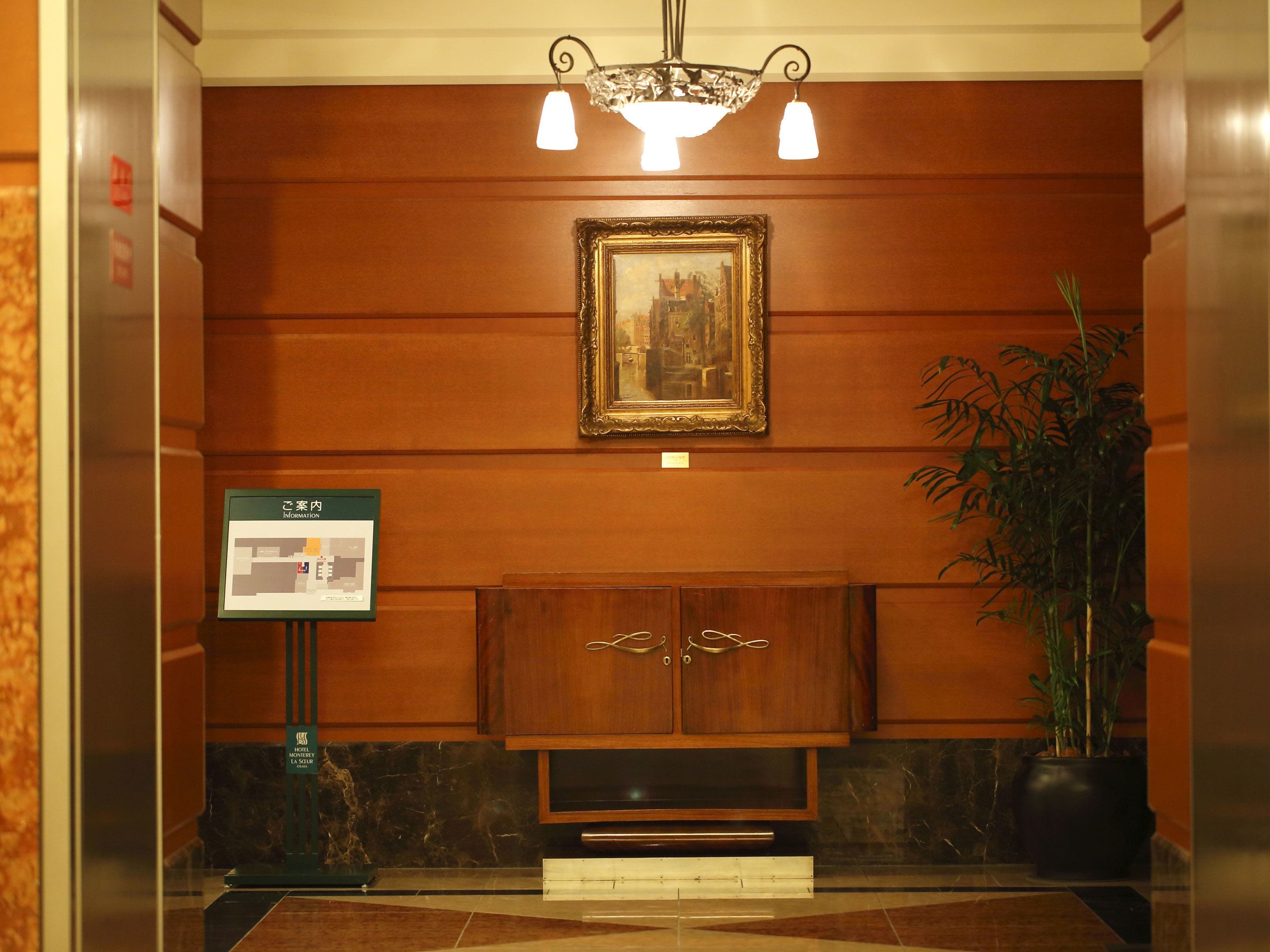 A hotel lobby with a wooden panel wall, a framed painting, a potted plant, and a digital information board.
