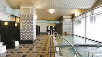 Una piscina cubierta pública con suelo de baldosas, una pileta con baranda metálica y techo de paneles de madera.