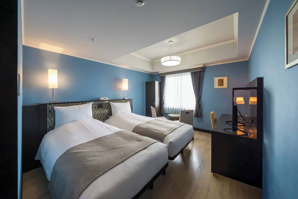 Une chambre d’hôtel avec deux lits, un bureau, une chaise, un miroir et une fenêtre avec des rideaux.