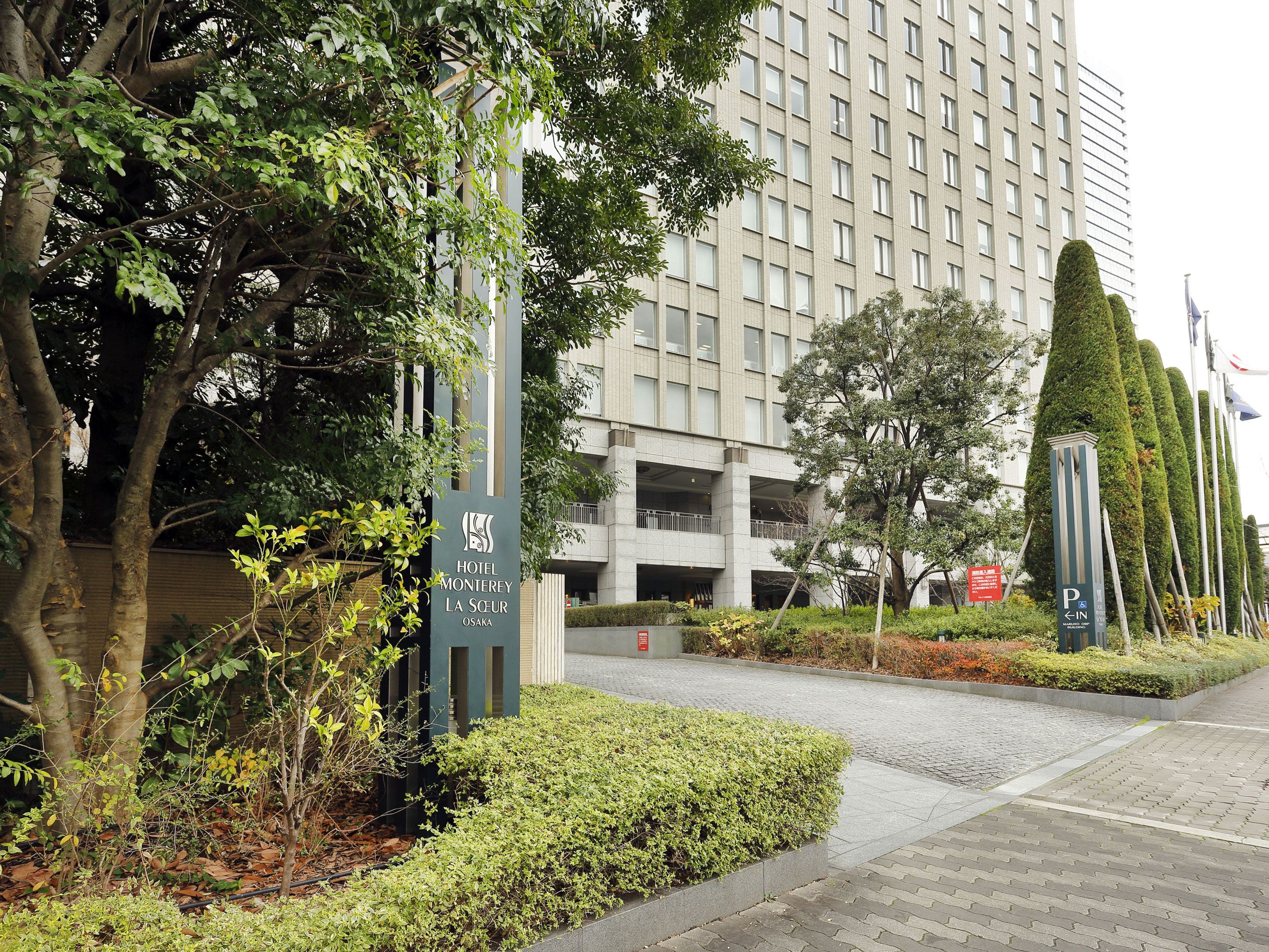 A modern hotel entrance with a sign for Hotel Monterey La Soeur in Osaka, surrounded by well-maintained greenery and a parking area.
