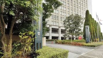 A modern hotel entrance with a sign for Hotel Monterey La Soeur in Osaka, surrounded by well-maintained greenery and a parking area.