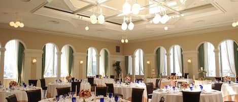 A spacious dining area with round tables, chandeliers, and large windows with curtains.