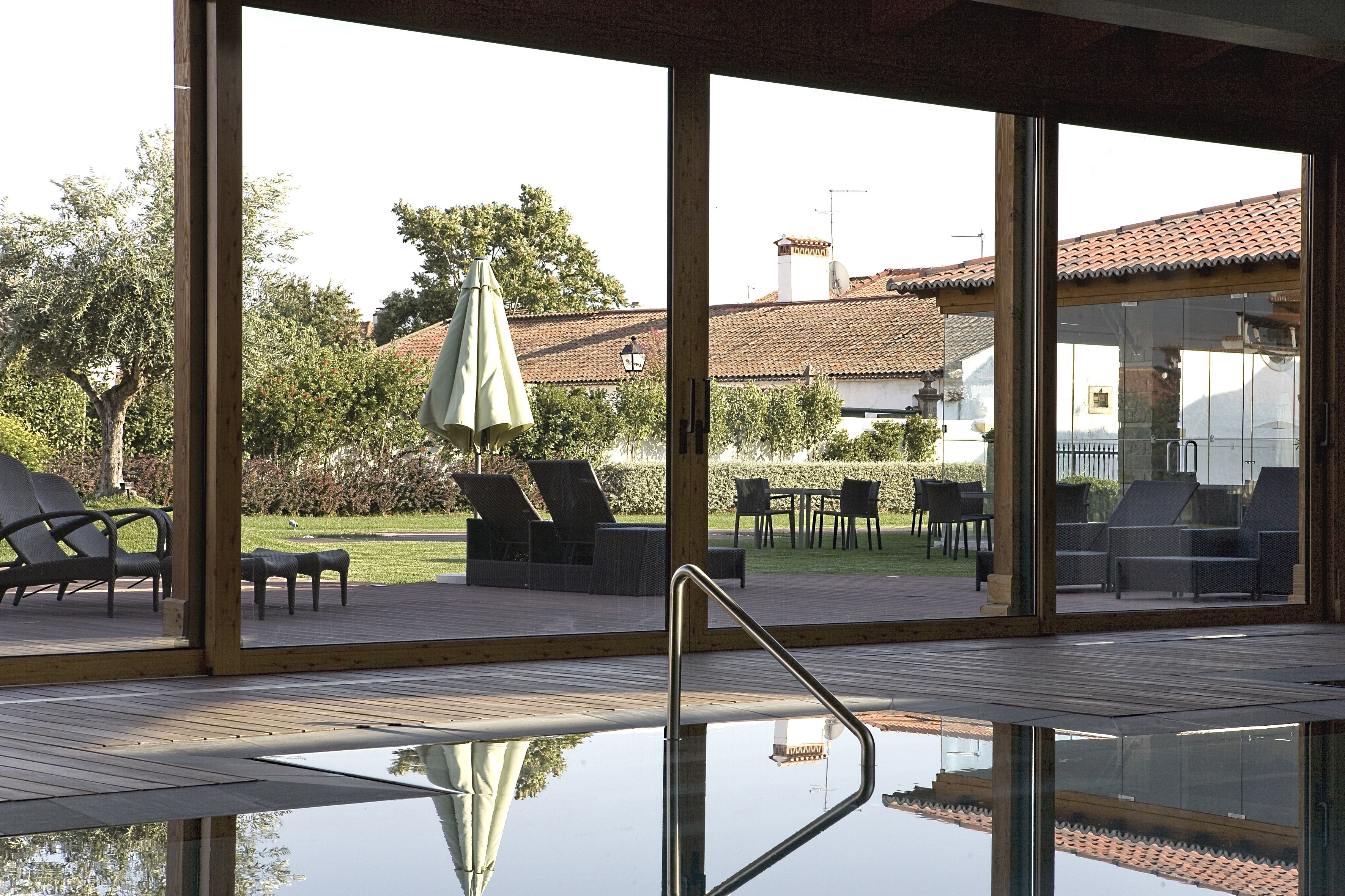 An indoor swimming pool with clear glass walls, lounge chairs, and umbrellas outside.