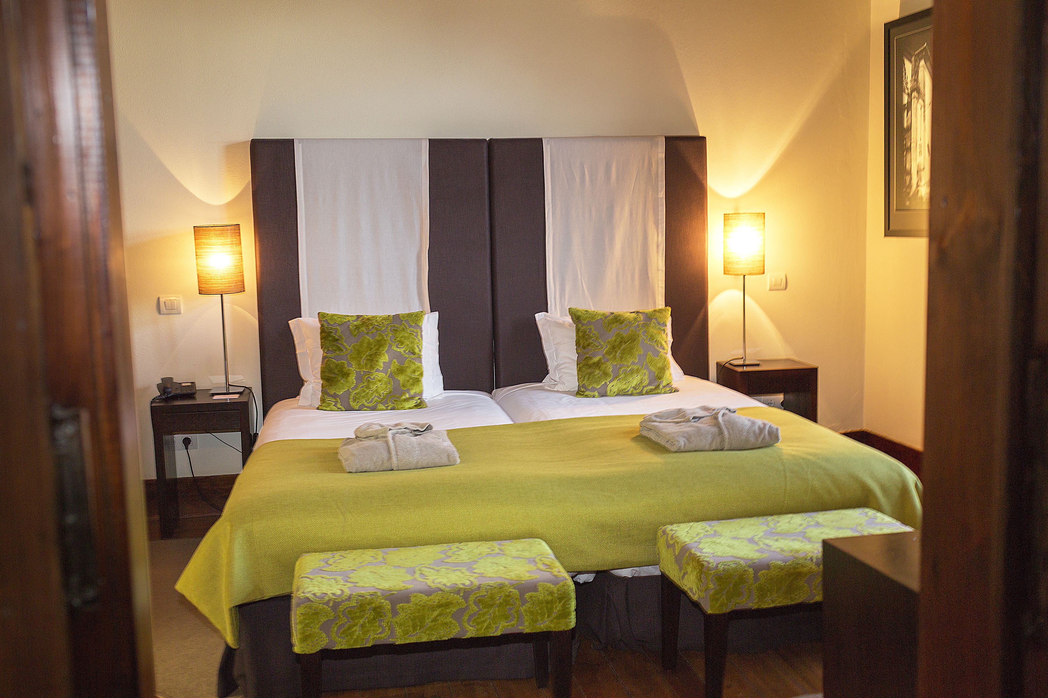 Una cama doble con cubrecama verde, dos almohadas y dos mesitas de noche con lámparas. Hay dos bancos iguales al pie de la cama.