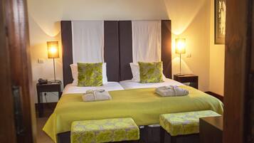 Una cama doble con cubrecama verde, dos almohadas y dos mesitas de noche con lámparas. Hay dos bancos iguales al pie de la cama.