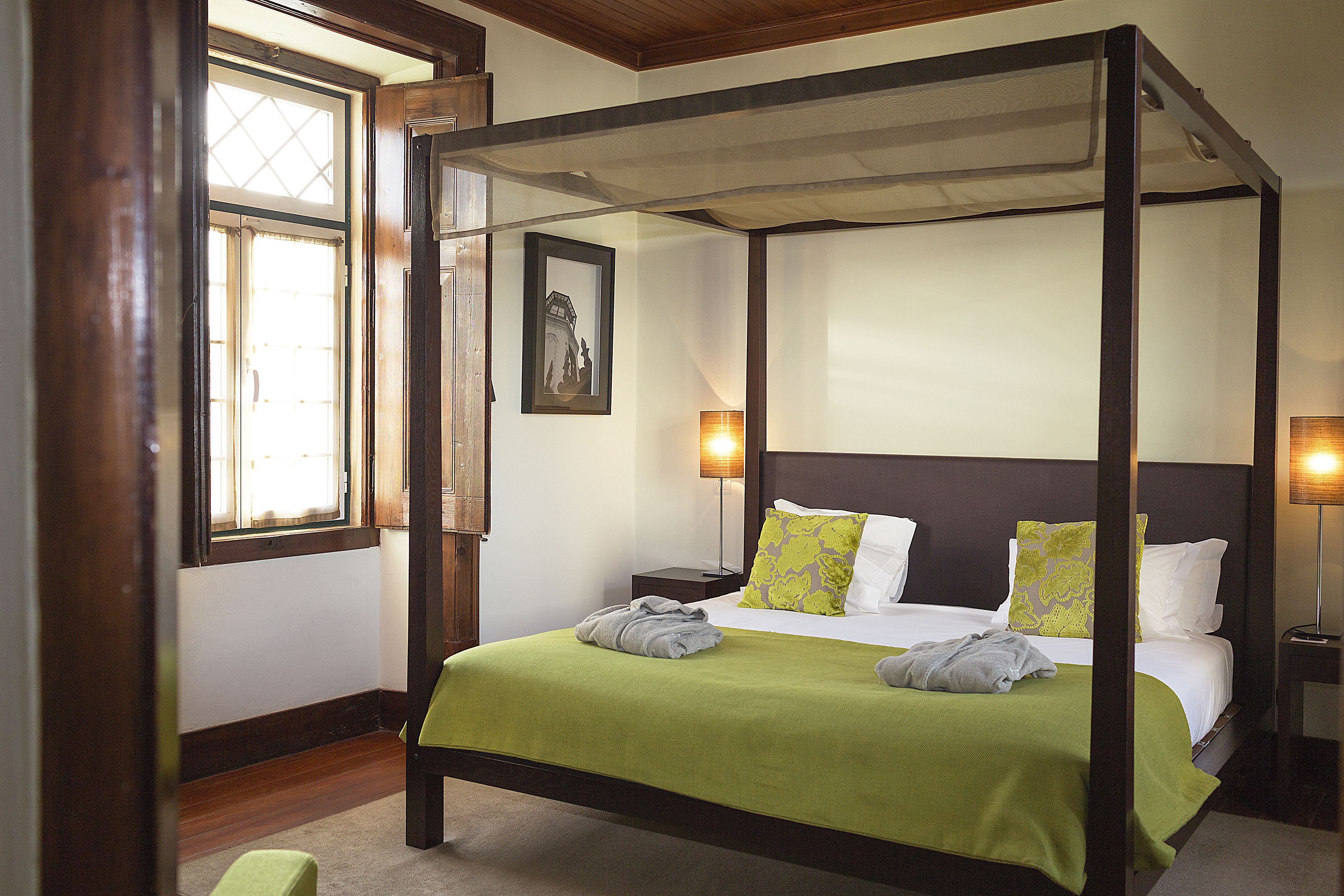 Een hemelbed met een groene plaid, twee bedlampjes en een ingelijst schilderij aan de muur.