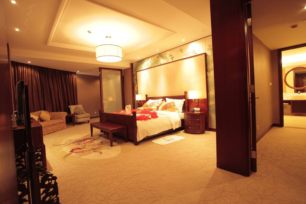 Una habitación de hotel con una cama grande, una zona de estar con un sofá y sillón, una mesita y un espejo.