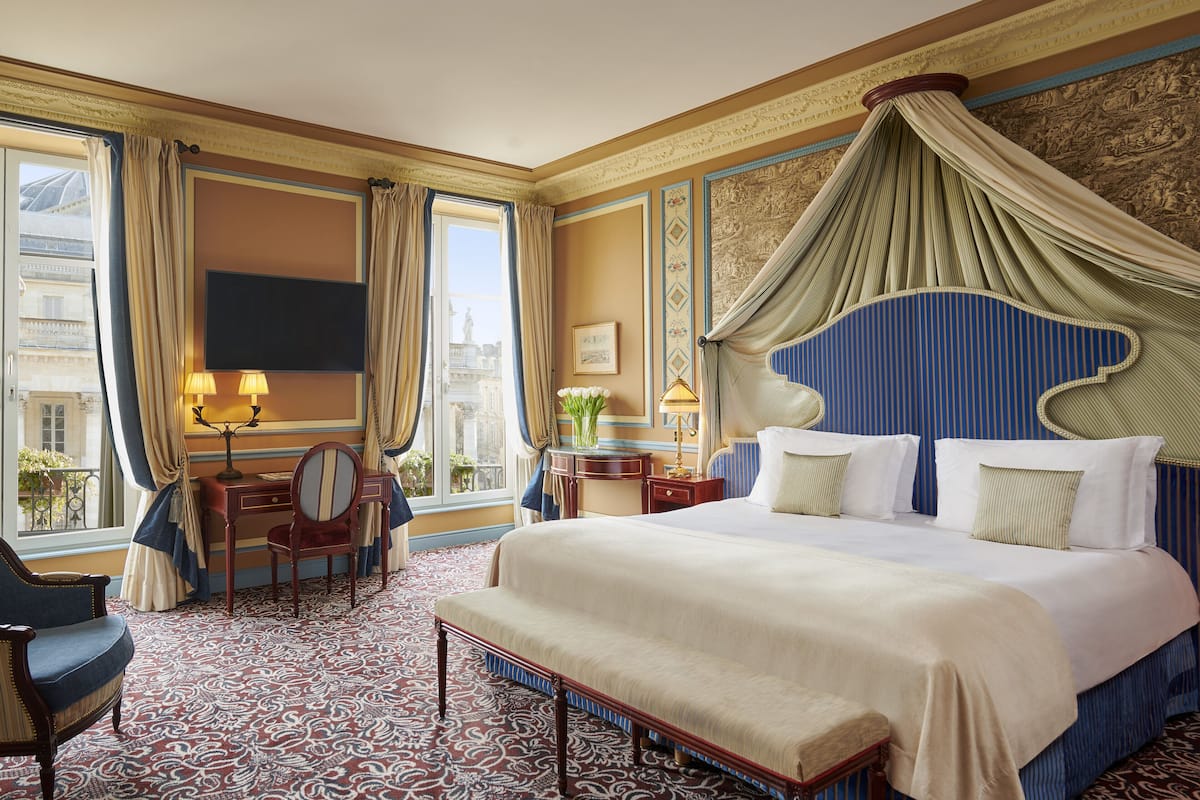 A hotel room with a large bed, a desk, a chair, a television, and a view of the city.