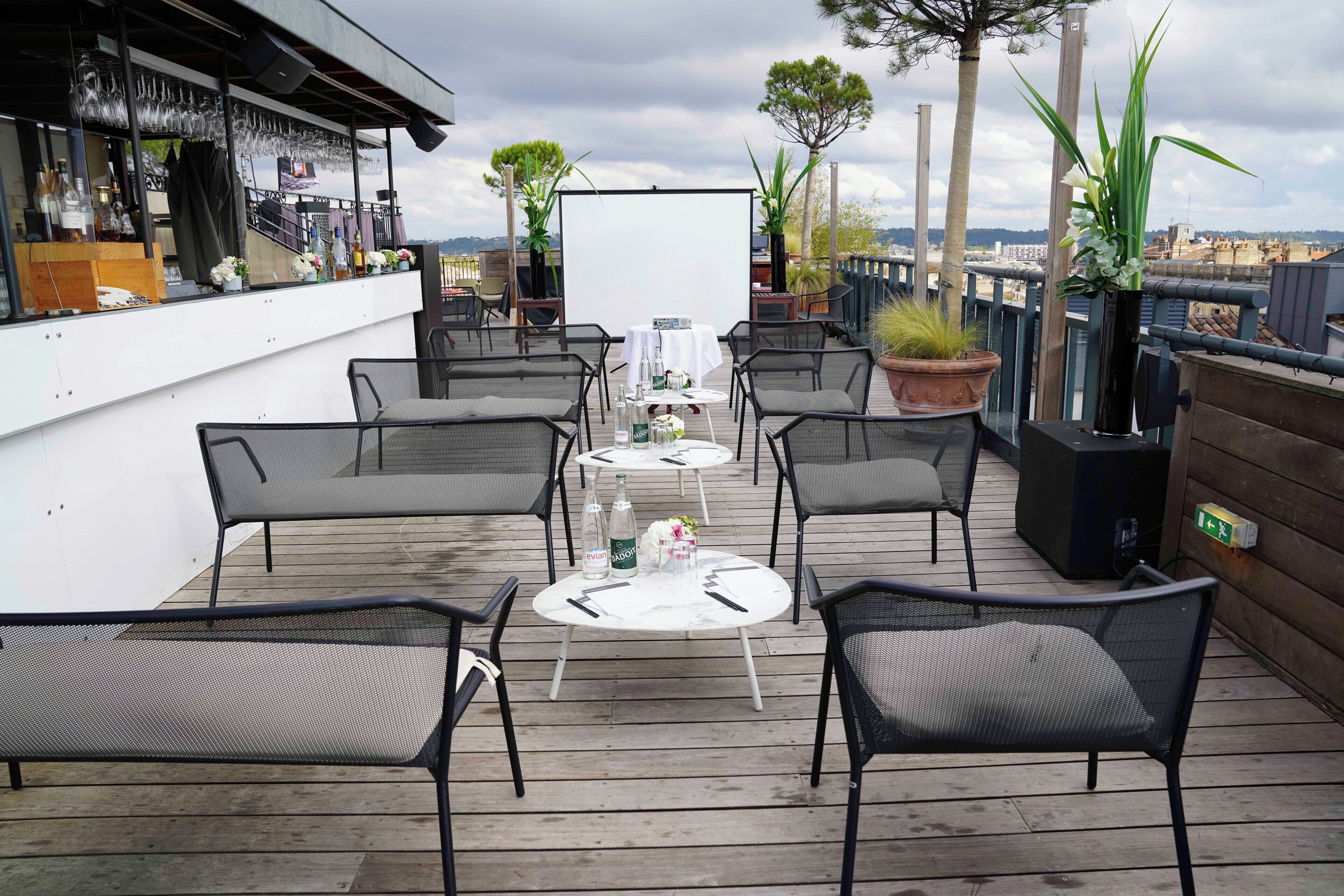 An outdoor patio with round tables and chairs, a bar area with glasses and bottles, and potted plants.