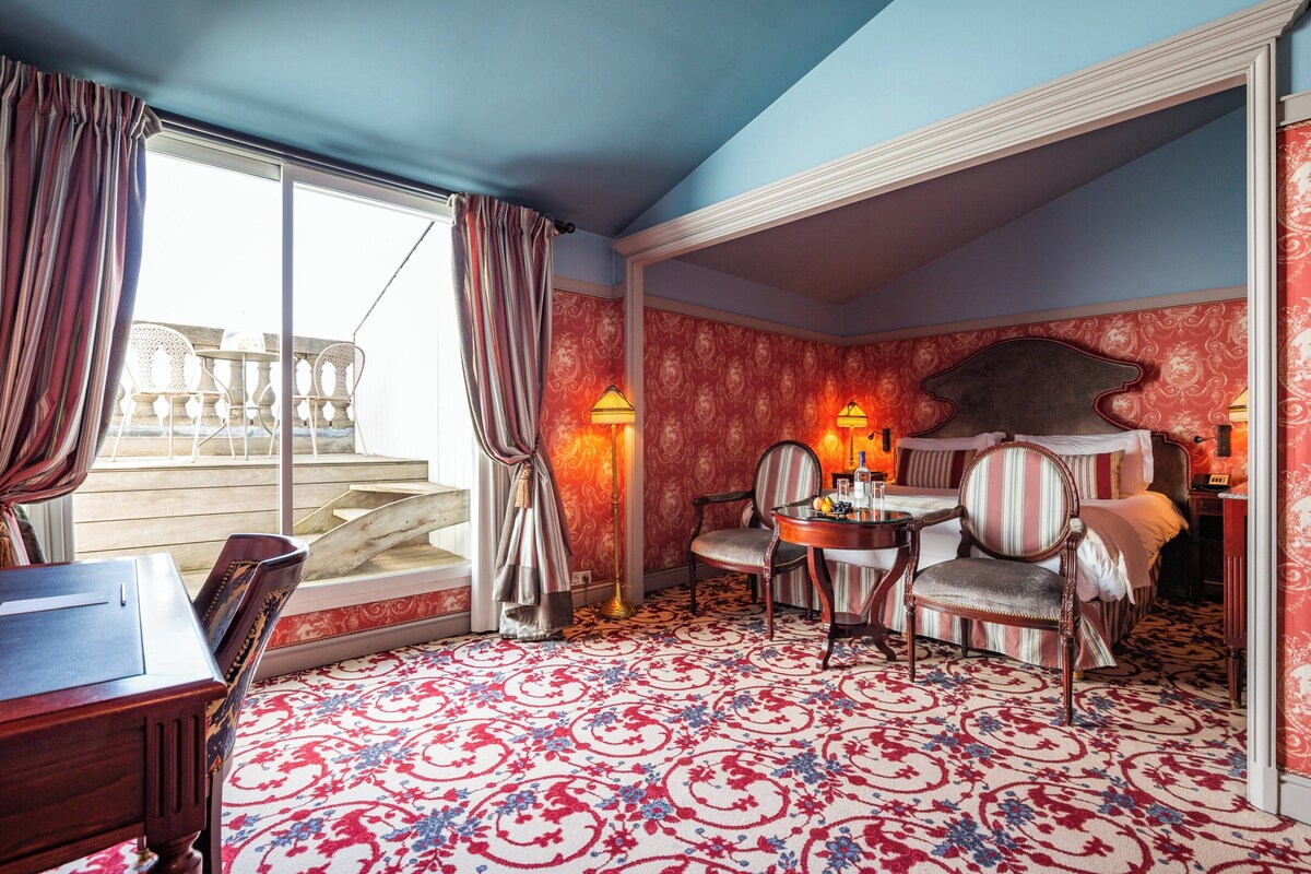 A room with a large window, a patterned carpet, a dining table with chairs, and a bed with a headboard.