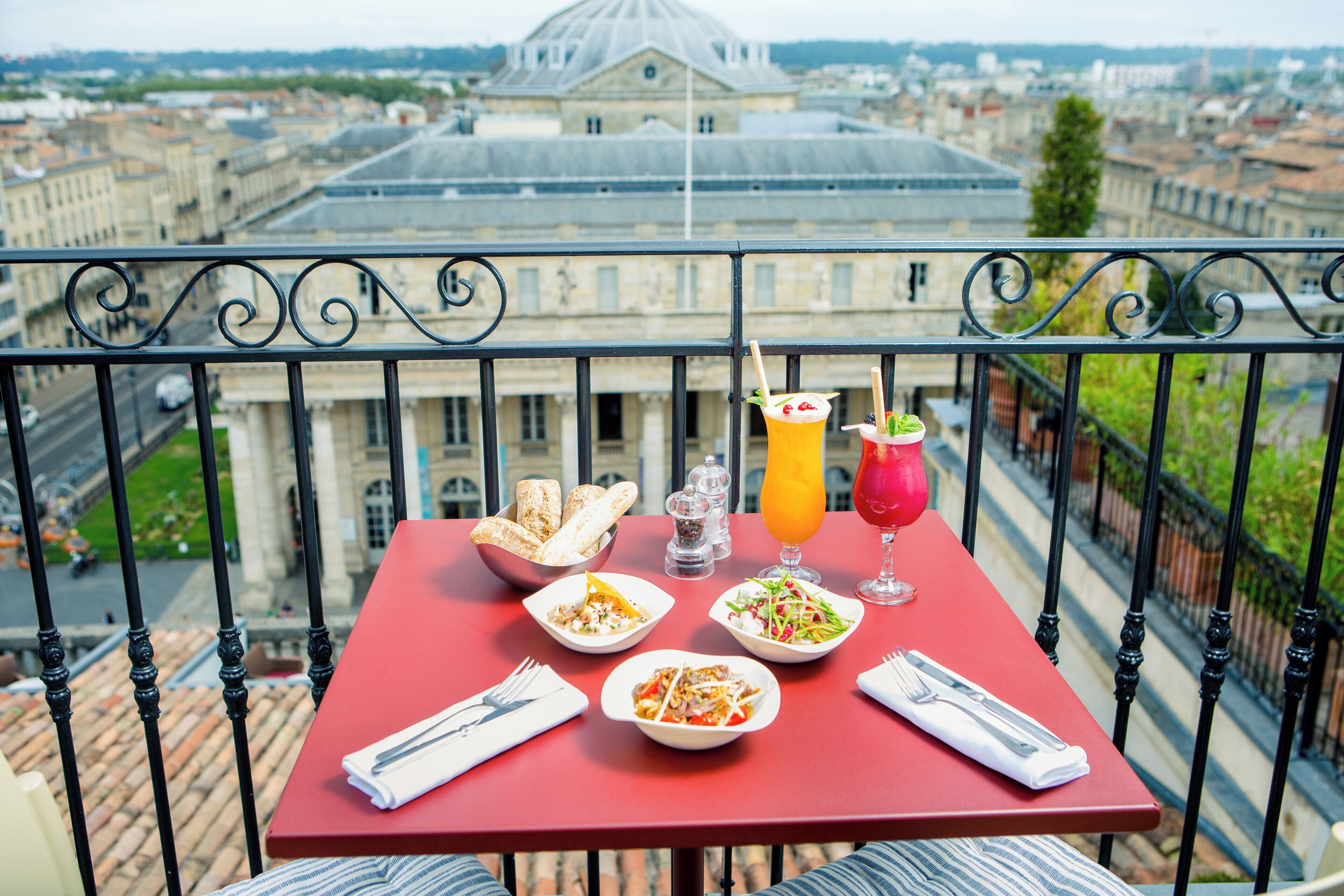 A balcony table with a red tablecloth, two plates of food, a glass of orange juice, a glass of red drink, and a bowl of breadsticks.