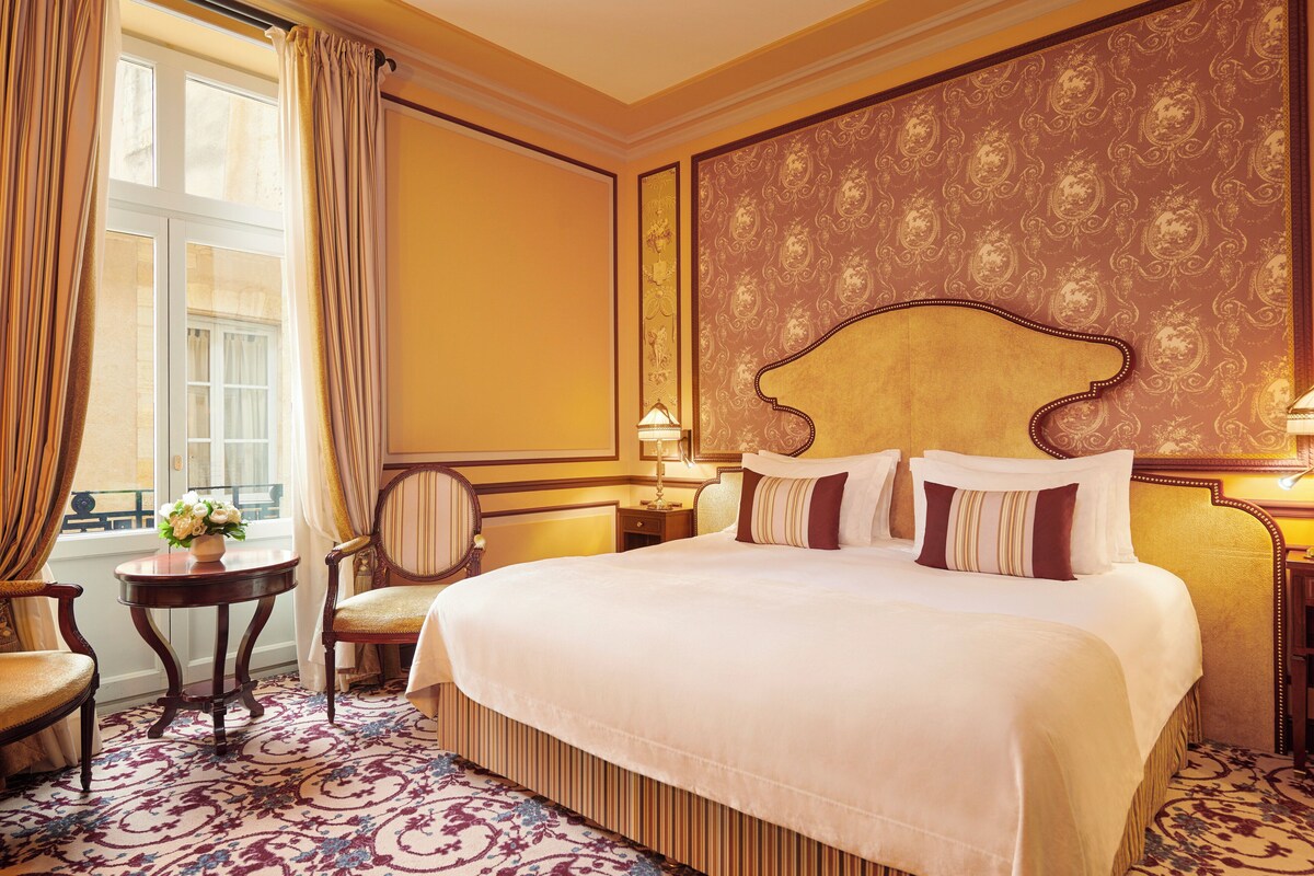 A hotel room with a large bed, a small round table with a lamp, a chair, and a window with curtains.