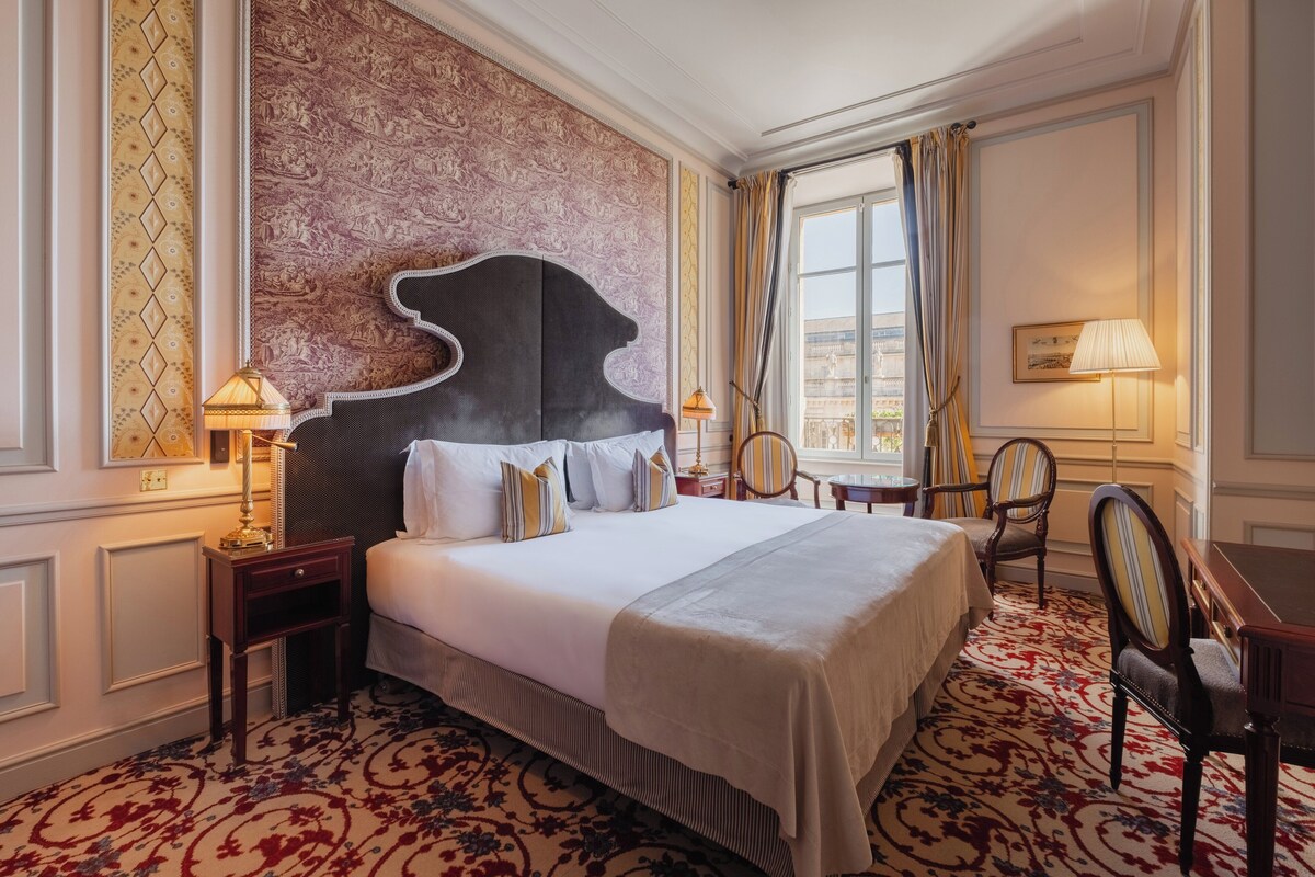 A hotel room with a large bed, a headboard, a nightstand, a chair, a desk, and a window with curtains.
