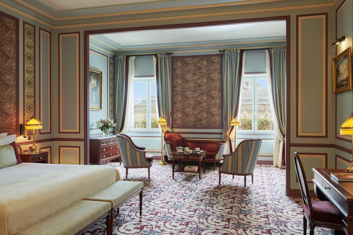 A hotel room with a large bed, a desk with a lamp, a chair, a small table with a vase of flowers, and a view of the city through the window.