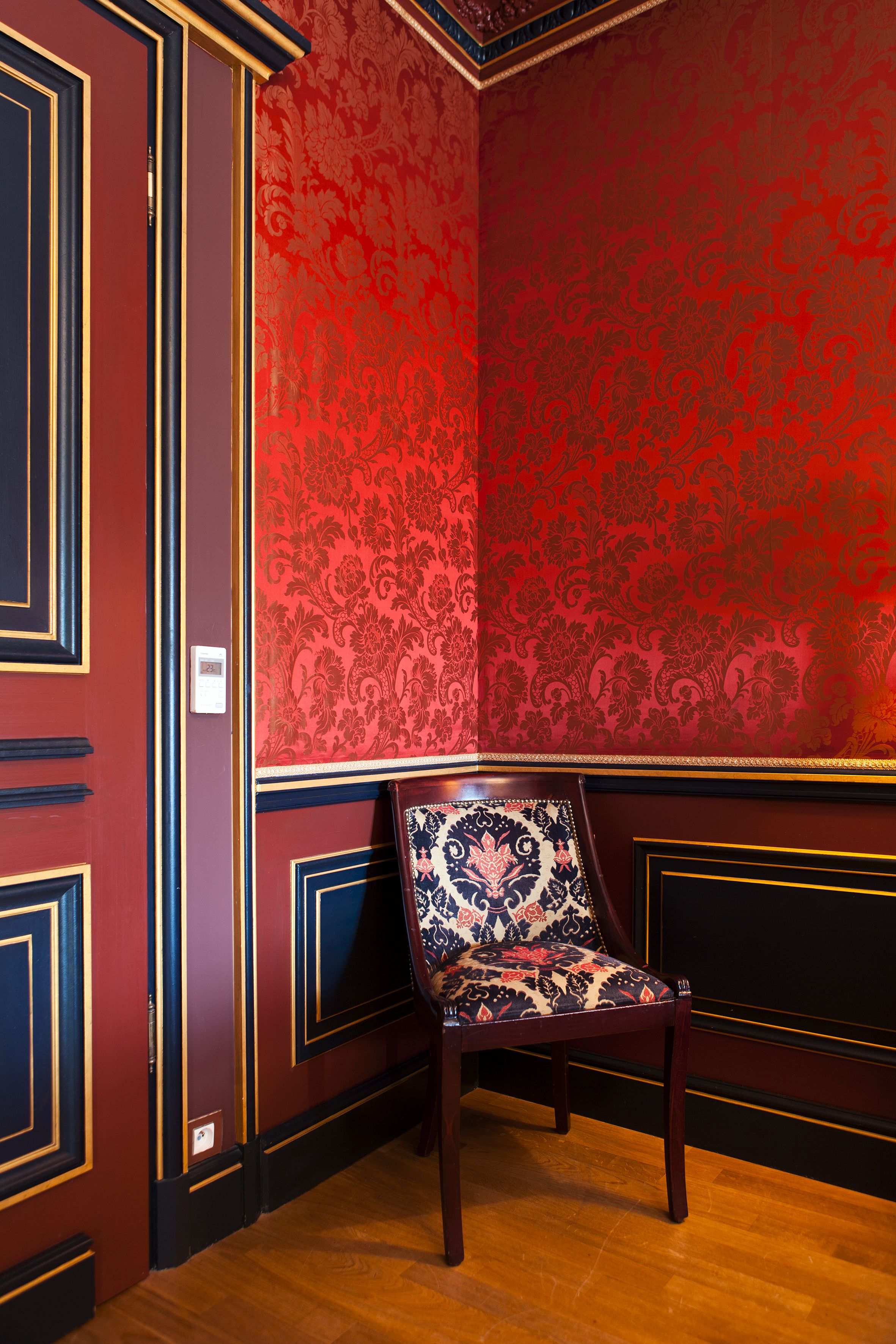 A corner room with red patterned wallpaper, a wooden chair, and dark wood paneling.