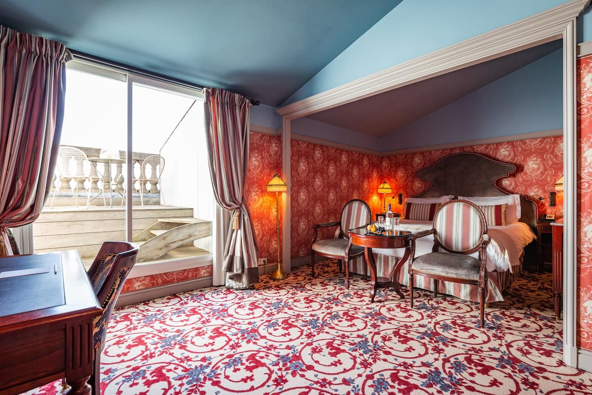 A room with a large window, a patterned carpet, a dining table with chairs, and a bed with a headboard.