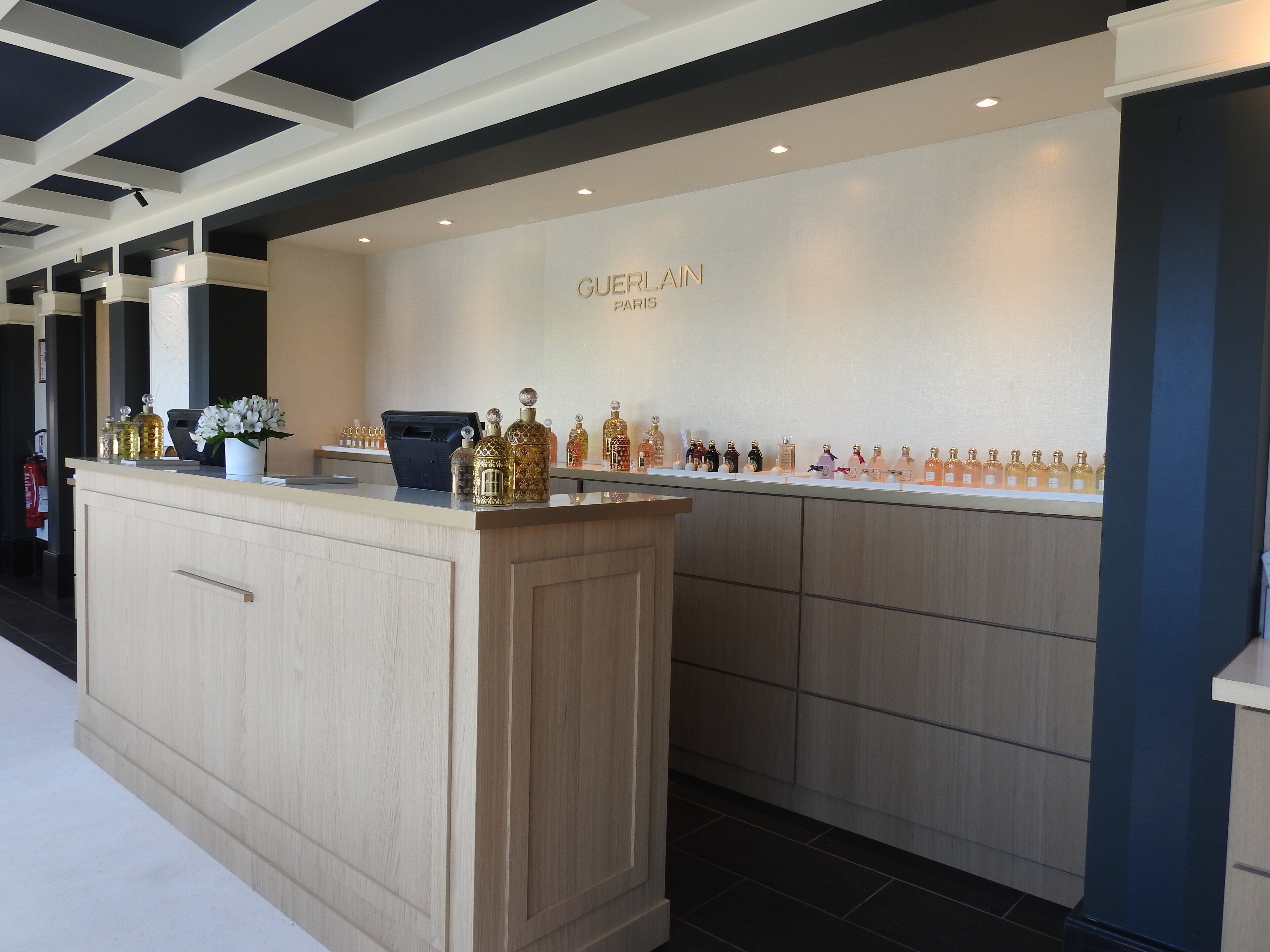Un área de recepción moderna en un hotel con un mostrador de madera clara, una estación de registro digital y una exhibición de frascos de perfume.