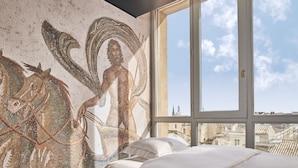 A room with a large window, a bed with white bedding, and a mosaic wall depicting a figure with a horse.