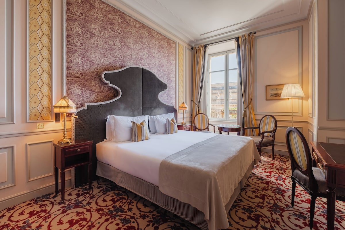 A hotel room with a large bed, a headboard, a nightstand, a chair, a desk, and a window with curtains.