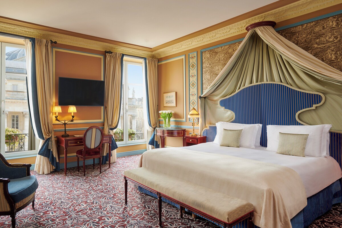 A hotel room with a large bed, a desk, a chair, a television, and a view of the city.