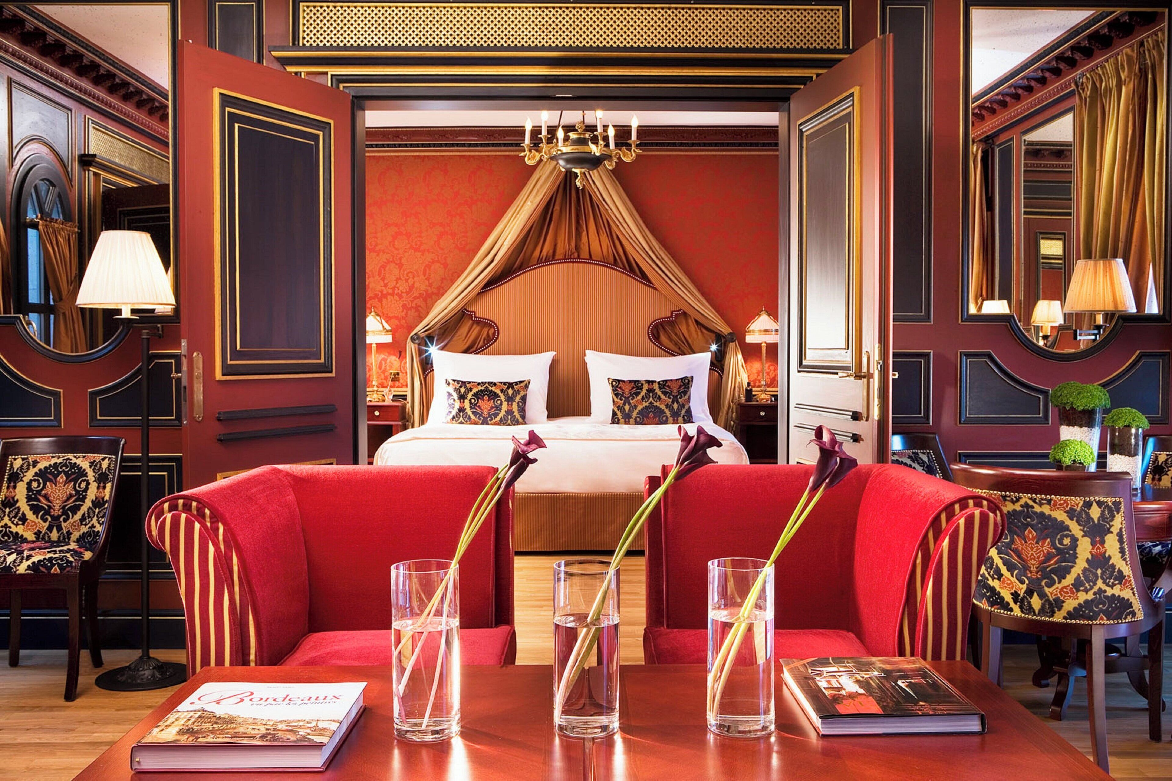 Une chambre d’hôtel luxueuse avec un grand lit, un canapé rouge et une table à manger sur laquelle se trouvent un livre et des verres d’eau.