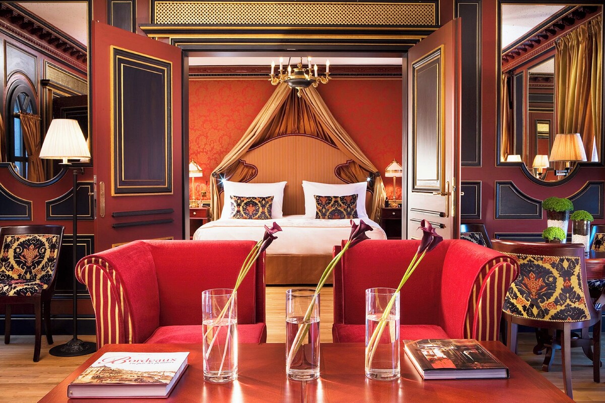 A luxurious hotel room with a large bed, red upholstered furniture, and a chandelier.