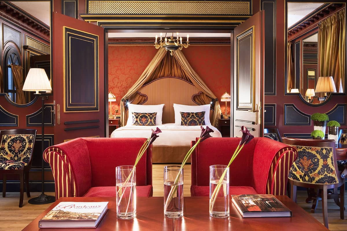 A luxurious hotel room with a large bed, red upholstered furniture, and a chandelier.