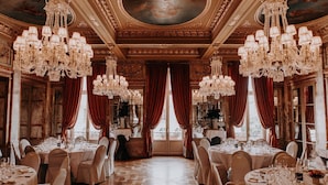 A luxurious dining room with ornate chandeliers, red curtains, and a polished wooden floor.