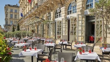 An outdoor dining area with tables and chairs, a building with large windows, and a flag.