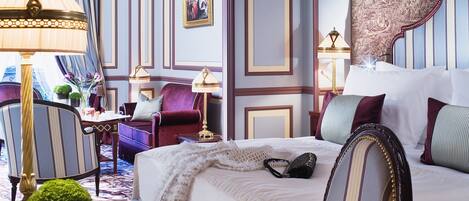 A well-appointed hotel room with a large bed, a sitting area with a chair and a small table, and a decorative lamp.