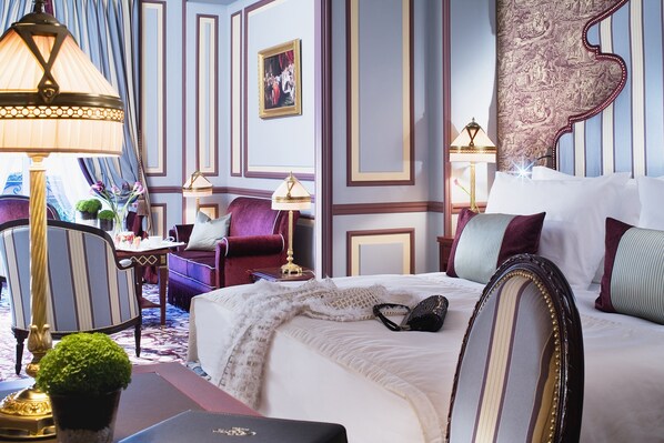 A well-appointed hotel room with a large bed, a sitting area with a chair and a small table, and a decorative lamp.