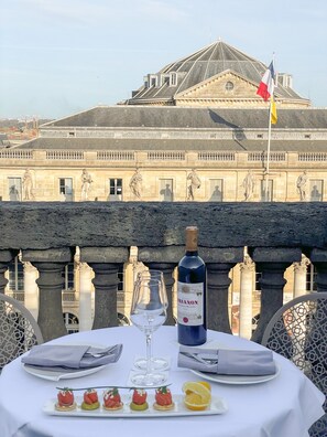 A table set with a bottle of wine, glasses, and hors d'oeuvres, overlooking a historic building with classical architecture and statues.