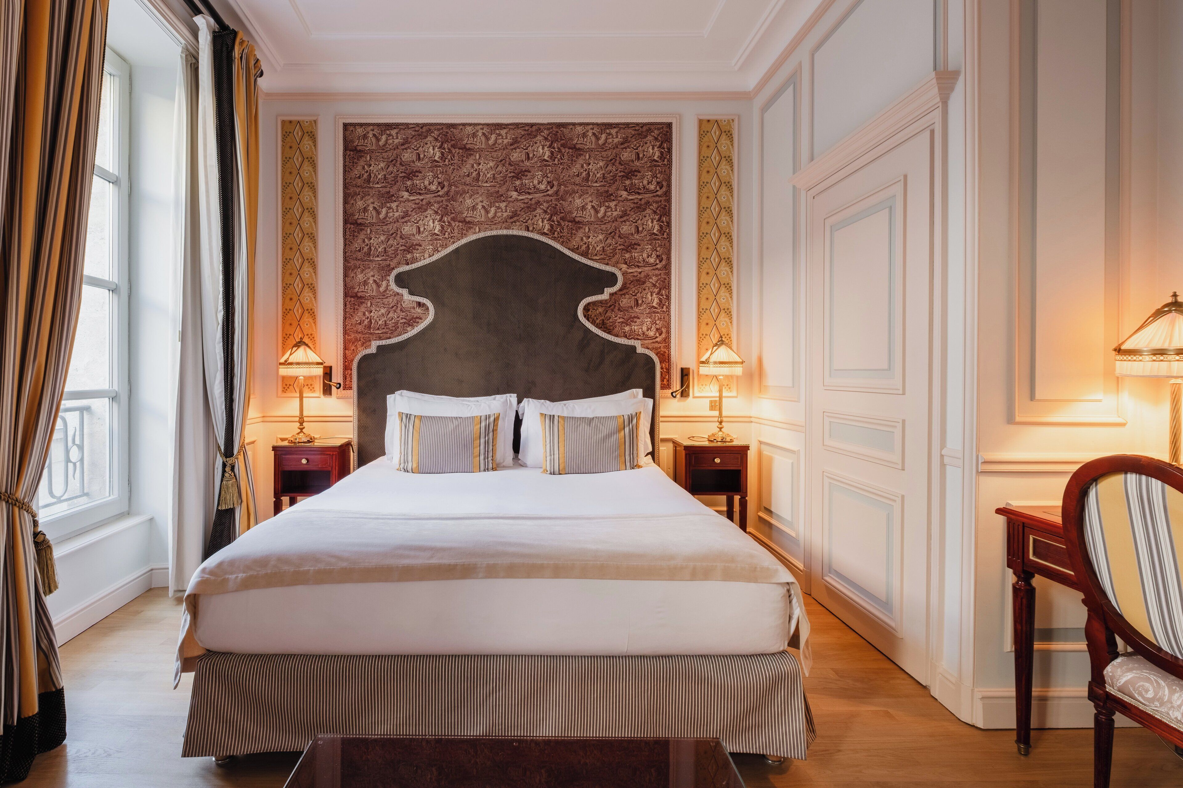 A large bed with a headboard, two bedside tables with lamps, a chair, and a patterned wall.
