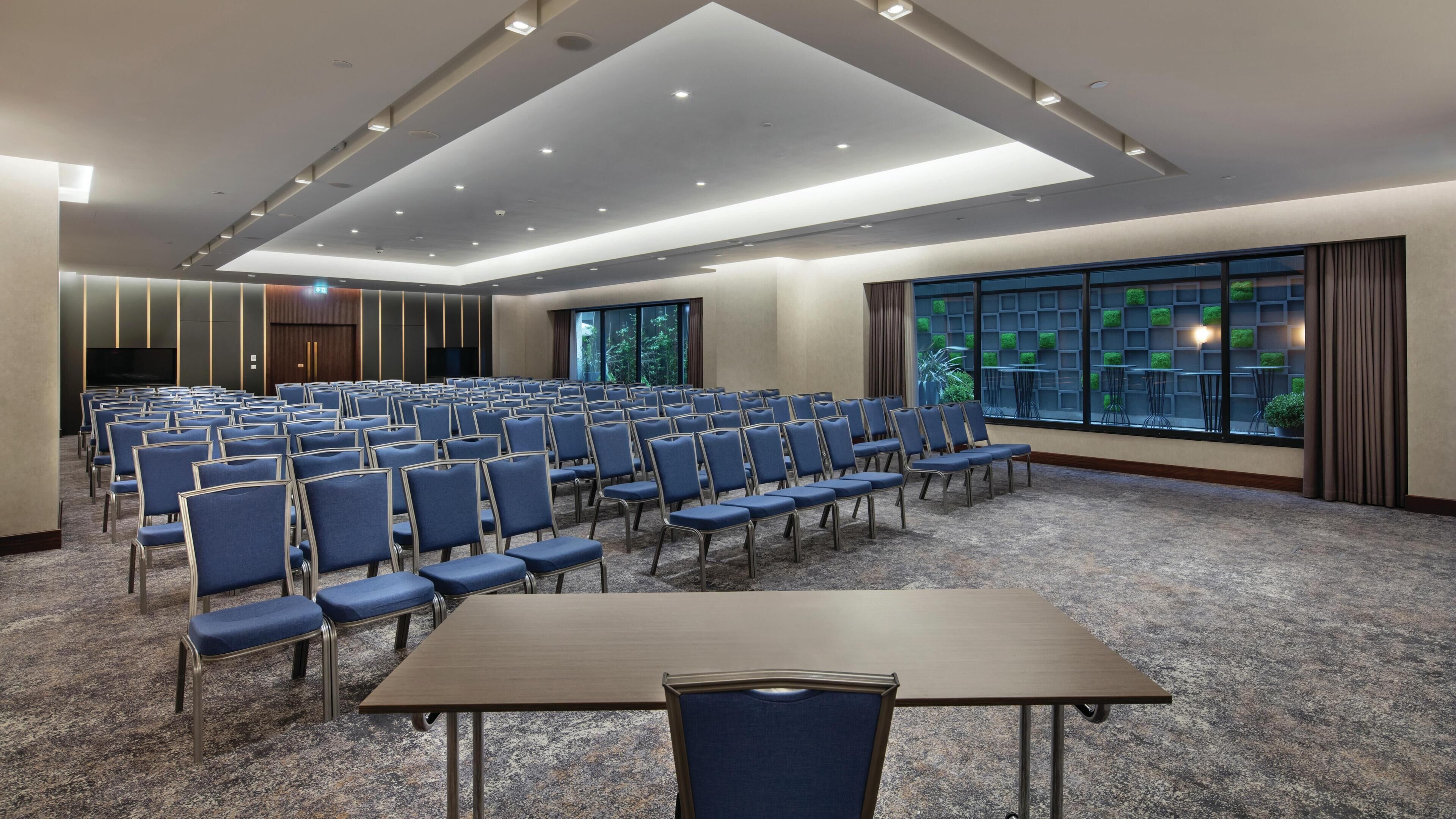 A conference room with rows of chairs, a large table, and a view of greenery through the windows.