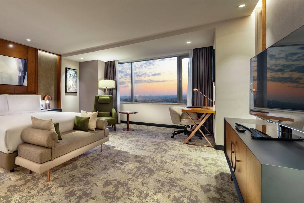 A hotel room with a bed, a sofa, a desk, a TV, and a large window with a sunset view.