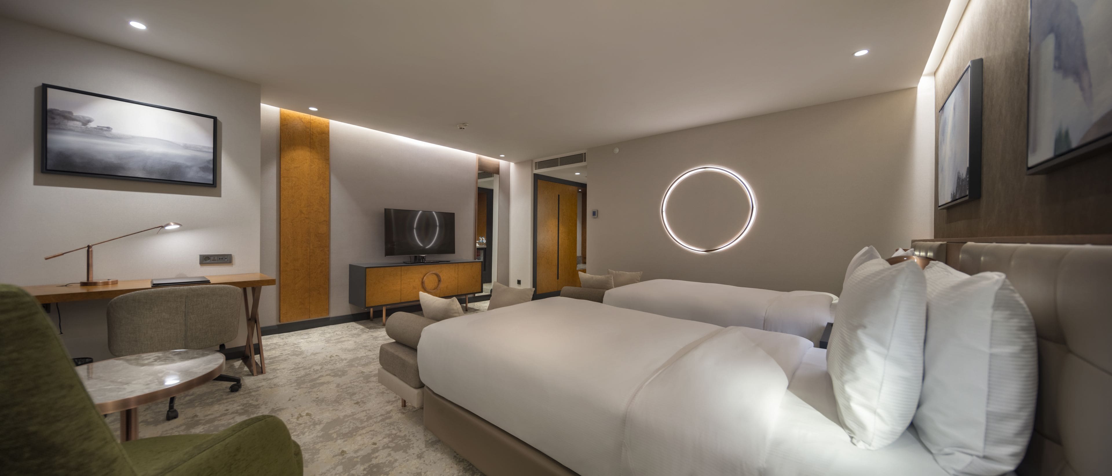 A modern hotel room with a large bed, a desk with a chair, a television, and a circular light fixture on the wall.