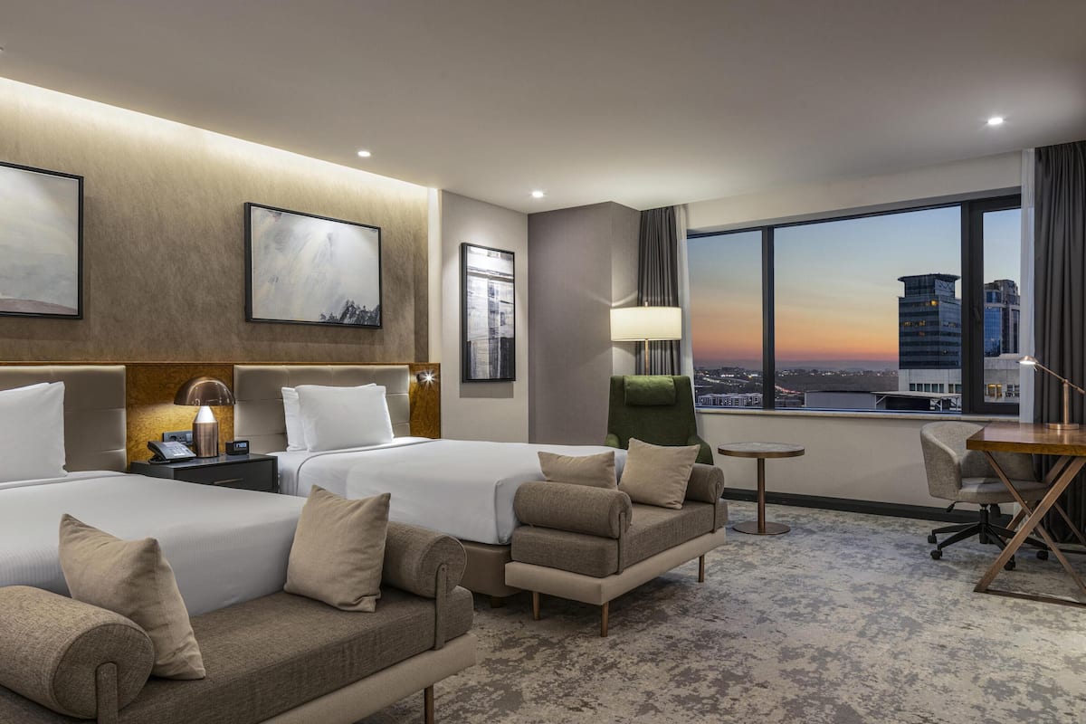A hotel room with two beds, a sofa, a desk, and a view of the cityscape.