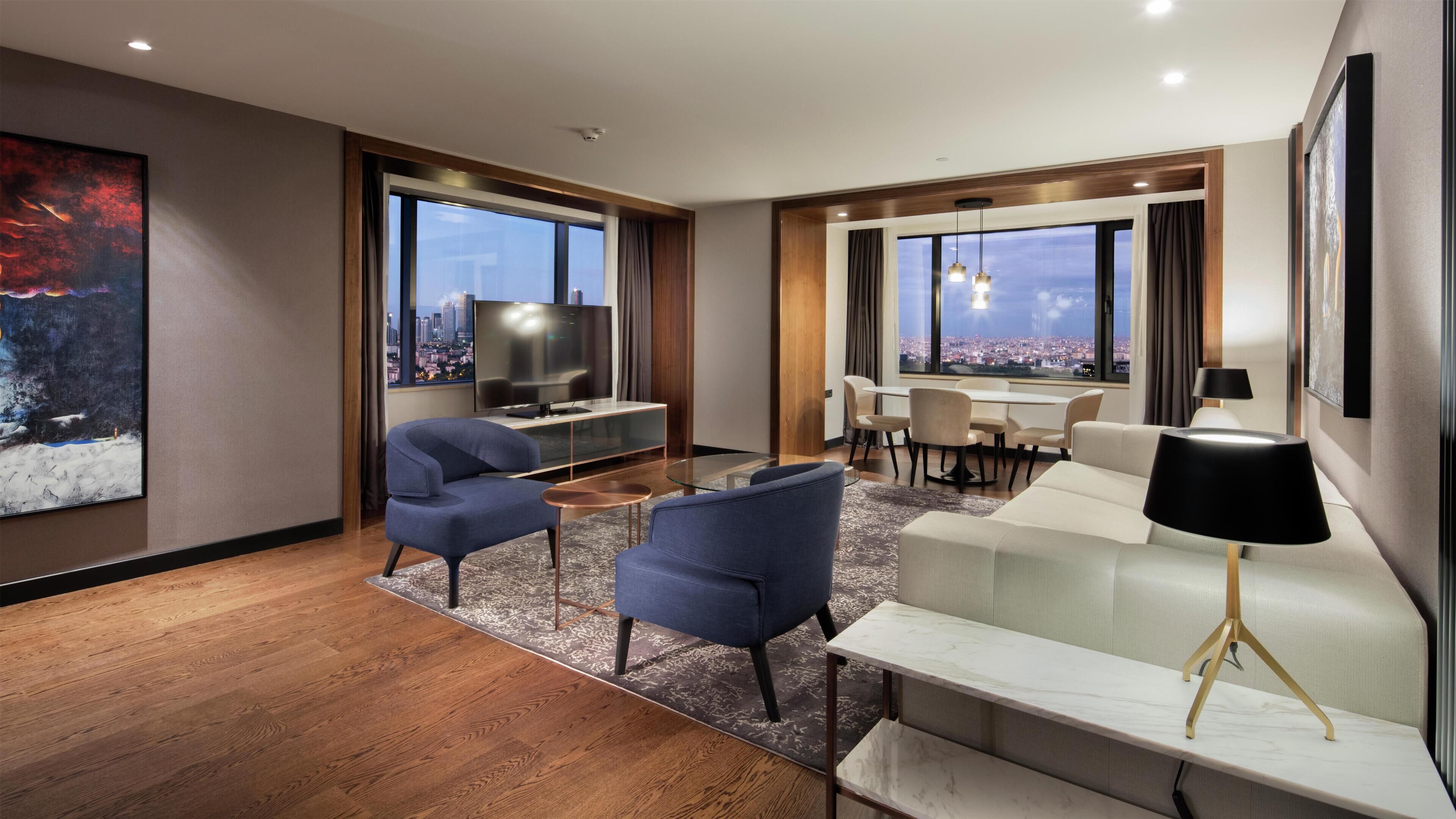 Habitación de hotel moderna con un ventanal grande, televisor de pantalla plana, sofá, mesa de centro y zona de comedor.