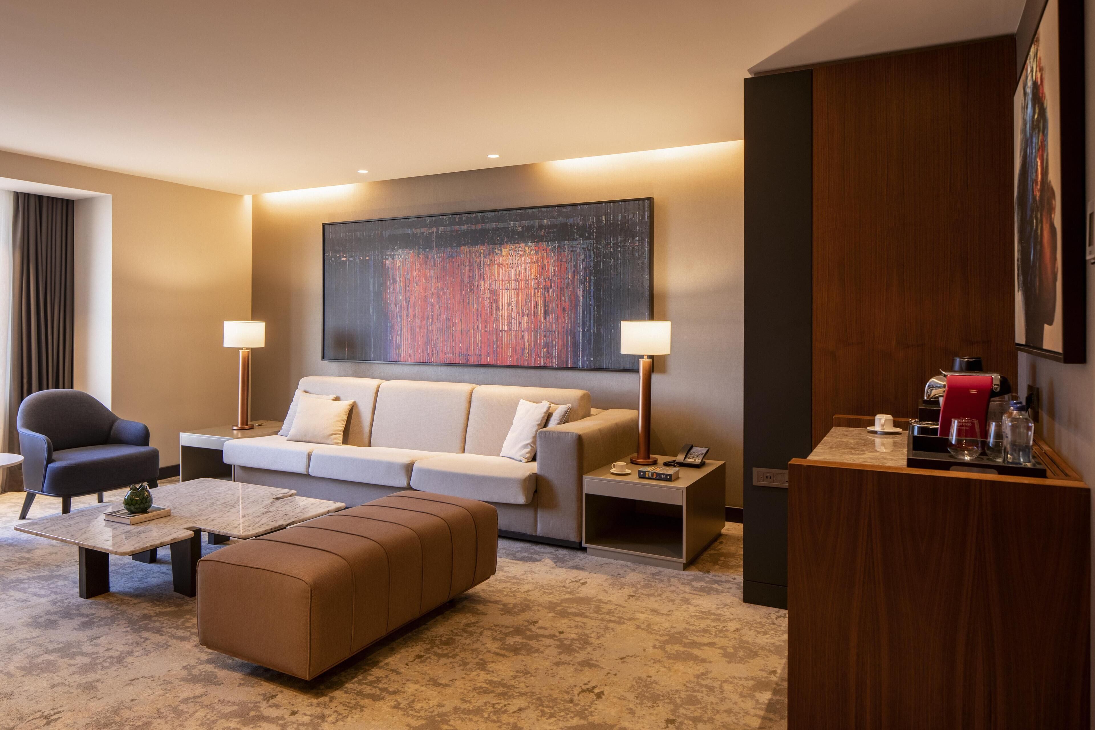 A modern hotel room with a sofa, armchairs, a coffee table, a side table, and a large abstract painting on the wall.